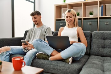 Obraz premium Young caucasian couple smiling happy using laptop and smartphone sitting on the sofa at home.
