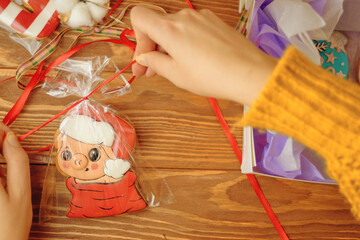 Person's hands tie red ribbon on bag of gingerbread in form of pig in Santa Claus hat. Cookies with colored icing in box on wooden table. Christmas gifts. New Year's atmosphere.