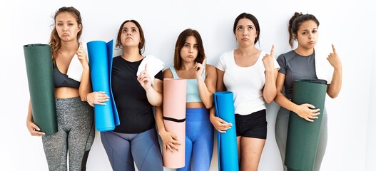 Obraz premium Group of women holding yoga mat standing over isolated background pointing up looking sad and upset, indicating direction with fingers, unhappy and depressed.