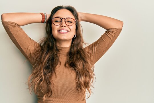 Young hispanic girl wearing casual clothes and glasses relaxing and stretching, arms and hands behind head and neck smiling happy