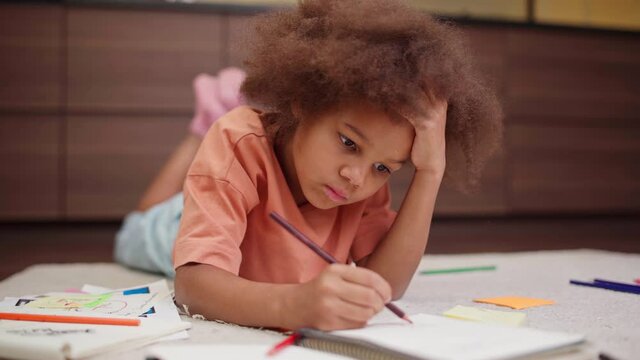 Portrait of cute biracial little girl lying on floor at home and drawing with pencils in notepad. Kid with brown eyes and curly hair looking at camera over homework or leisure activity