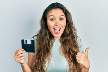 Young hispanic girl holding floppy disk pointing thumb up to the side smiling happy with open mouth