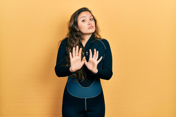 Young hispanic girl wearing diver neoprene uniform moving away hands palms showing refusal and...