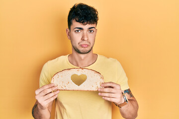 Young hispanic man holding bread with heart shape over eye depressed and worry for distress, crying angry and afraid. sad expression.