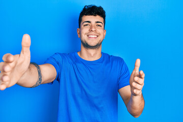 Young hispanic man wearing casual blue t shirt looking at the camera smiling with open arms for hug. cheerful expression embracing happiness.