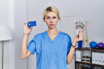 Young physiotherapist woman holding credit card and dollars banknotes skeptic and nervous, frowning...
