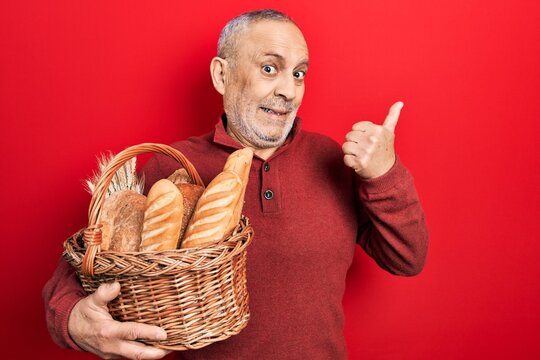 Handsome mature man holding wicker basket with bread pointing thumb up to the side smiling happy with open mouth