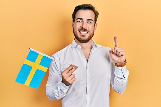 Handsome caucasian man with beard holding sweden flag smiling with an idea or question pointing finger with happy face, number one
