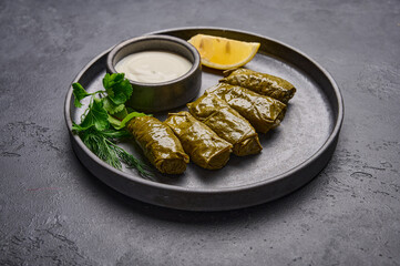 Middle Eastern dish dolma or sarma, with parsley, saucepan with sour cream and lemon in black plate on dark wood background. Close up