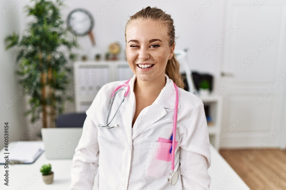 Poster young caucasian woman wearing doctor uniform and stethoscope at the clinic winking looking at the ca