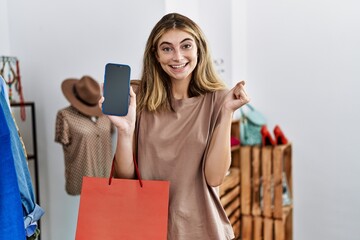 Young blonde woman holding shopping bags showing smartphone screen screaming proud, celebrating victory and success very excited with raised arm
