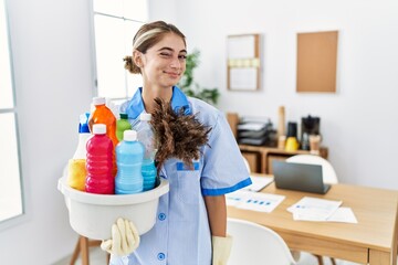 Young blonde woman wearing cleaner uniform holding cleaning products winking looking at the camera...