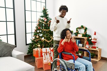 Young interracial couple with woman sitting on wheelchair by christmas tree pointing fingers to camera with happy and funny face. good energy and vibes.