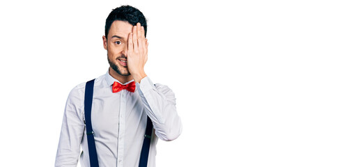 Hispanic man with beard wearing hipster look with bow tie and suspenders covering one eye with hand, confident smile on face and surprise emotion.