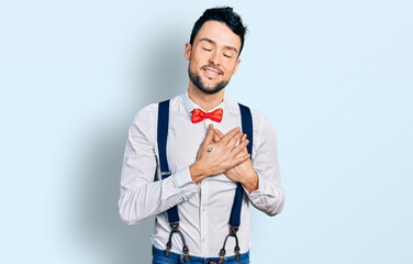 Hispanic man with beard wearing hipster look with bow tie and suspenders smiling with hands on...
