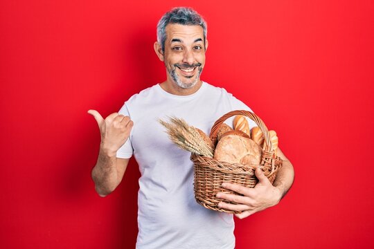 Handsome middle age man with grey hair holding wicker basket with bread pointing thumb up to the side smiling happy with open mouth