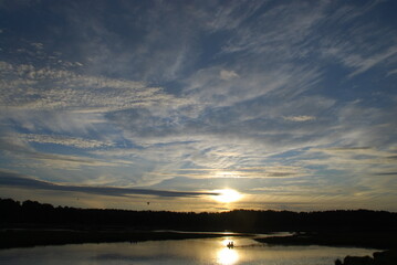 Sunset over the forest. Above the dark wall of the forest and the lake in white clouds, the sun is setting fancifully illuminating everything around. Sun rays are reflected from the surface of water.