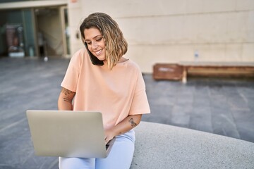 Young hispanic woman talking on the smartphone using laptop at street