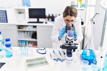 Young hispanic woman wearing scientist uniform using microscope at laboratory