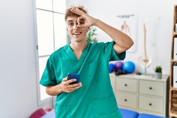 Young caucasian physio man using smartphone at medical clinic smiling happy doing ok sign with hand on eye looking through fingers