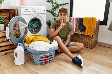 Young caucasian man putting dirty laundry into washing machine asking to be quiet with finger on lips. silence and secret concept.