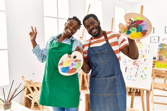 African american painter couple smiling happy holding paintbrush and palette at art studio.