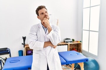 Handsome young man working at pain recovery clinic with hand on chin thinking about question, pensive expression. smiling with thoughtful face. doubt concept.