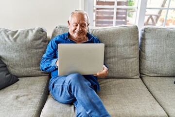 Senior man with grey hair sitting on the sofa at the living room of his house using computer laptop