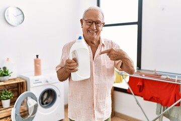 Senior man holding detergent bottle at laundry room smiling happy pointing with hand and finger