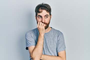 Caucasian man with beard wearing casual grey t shirt looking stressed and nervous with hands on mouth biting nails. anxiety problem.