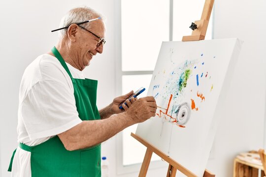 Senior grey-haired artist man smiling happy and using smartphone painting at art studio.