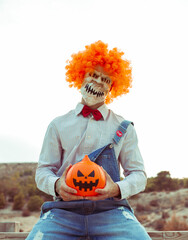 Payaso diabólico, sentado, sujetando una calabaza de Halloween con fondo blanco con montañas.