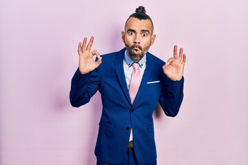 Young hispanic man wearing business suit and tie looking surprised and shocked doing ok approval symbol with fingers. crazy expression