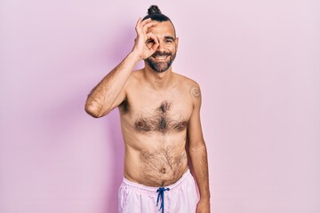 Fototapeta premium Young hispanic man shirtless wearing swimsuit doing ok gesture with hand smiling, eye looking through fingers with happy face.