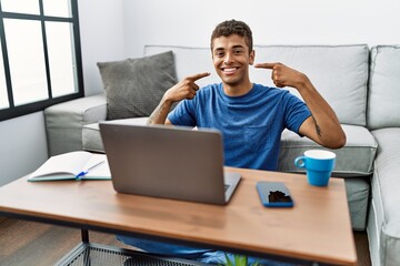 Young handsome hispanic man using laptop sitting on the floor smiling cheerful showing and pointing with fingers teeth and mouth. dental health concept.