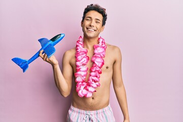 Young handsome man wearing swimsuit and hawaiian lei holding airplane toy looking positive and...