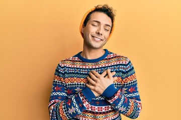 Fototapeta premium Young handsome man wearing wool hat and colorful sweater smiling with hands on chest with closed eyes and grateful gesture on face. health concept.