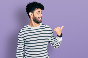 Young arab man with beard wearing casual striped sweater smiling with happy face looking and pointing to the side with thumb up.