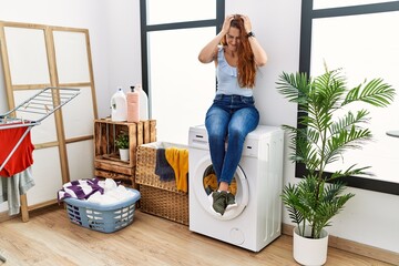 Young redhead woman doing laundry sitting on washing machine suffering from headache desperate and...
