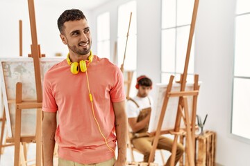 Young hispanic man at art studio looking away to side with smile on face, natural expression. laughing confident.