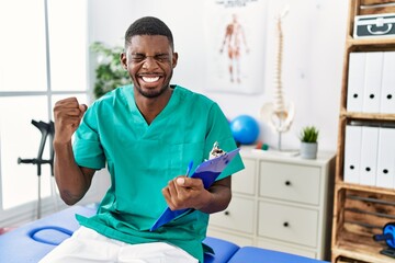 Young african american man working at pain recovery clinic very happy and excited doing winner gesture with arms raised, smiling and screaming for success. celebration concept.