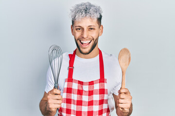Naklejka premium Young hispanic man with modern dyed hair wearing cook apron holding baker whisk and spoon smiling and laughing hard out loud because funny crazy joke.