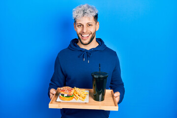 Young hispanic man with modern dyed hair eating a tasty classic burger with fries and soda smiling and laughing hard out loud because funny crazy joke.