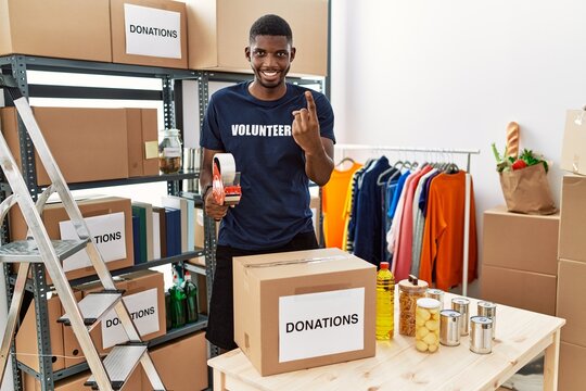 Young african american volunteer man packing donations box for charity beckoning come here gesture with hand inviting welcoming happy and smiling