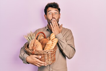 Handsome man with beard holding wicker basket with bread covering mouth with hand, shocked and...