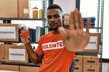 Young african man wearing volunteer t shirt at donations stand holding heart with open hand doing...