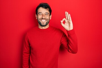 Handsome man with beard wearing casual red sweater smiling positive doing ok sign with hand and fingers. successful expression.