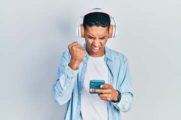 Young african american guy using smartphone wearing headphones annoyed and frustrated shouting with anger, yelling crazy with anger and hand raised