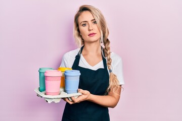 Beautiful young blonde woman wearing waitress apron holding take away cup of coffees relaxed with serious expression on face. simple and natural looking at the camera.