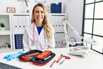 Young beautiful doctor woman with reflex hammer and medical instruments sticking tongue out happy...
